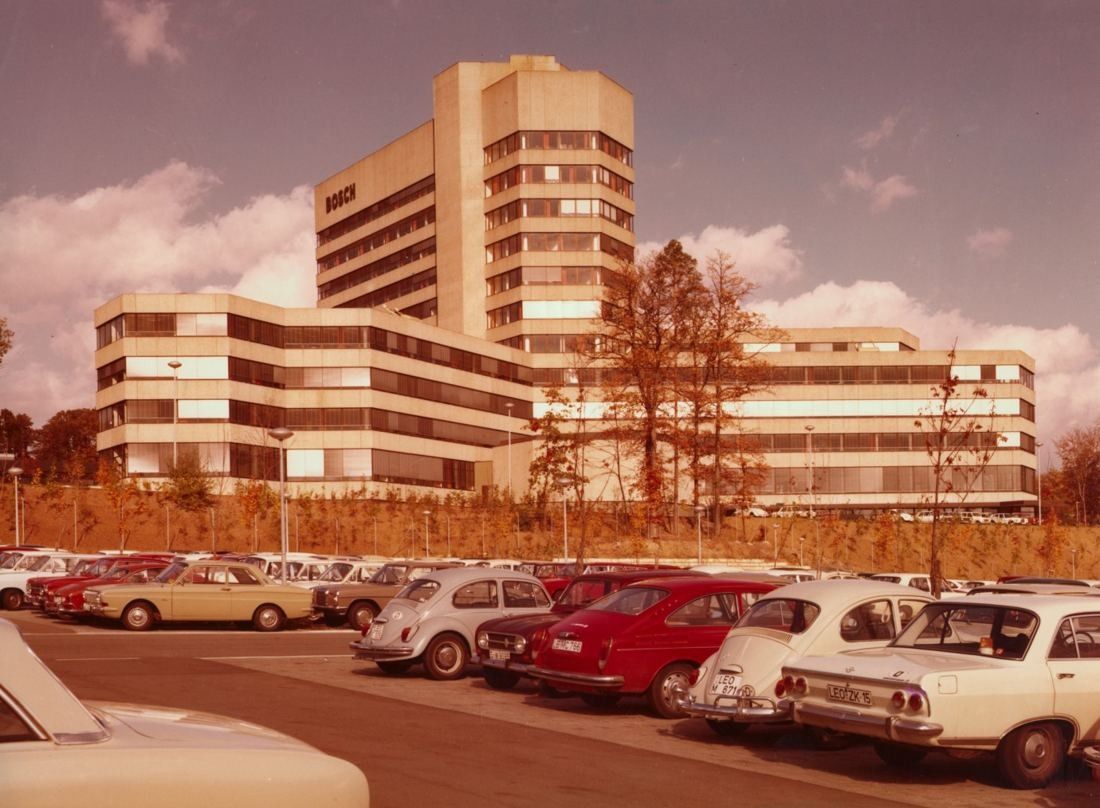 Colour photo of a tiered office complex with a tower block, parked cars in front