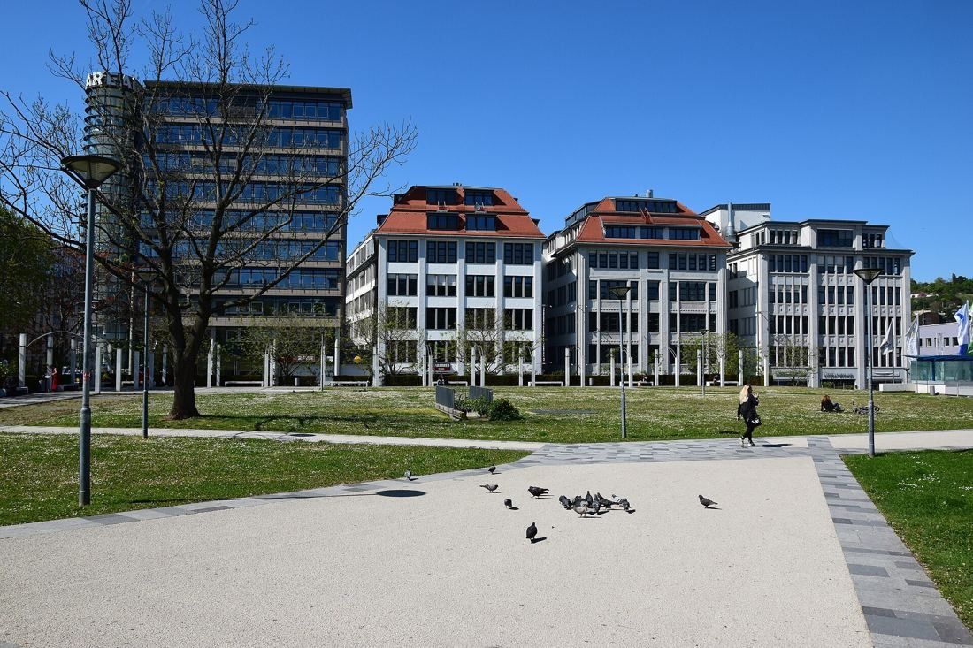 Restored historic white buildings with red roofs beyond a lawn and a paved square, modern tower at left
