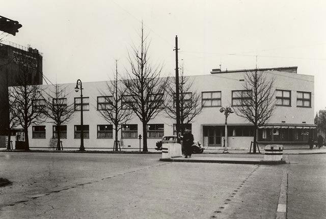 A black-and-white photo of a low, two-storey office building on a tree-lined street corner