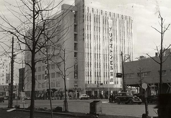 A black-and-white photo of a tall, light-coloured office building with a vertical sign, beside bare trees