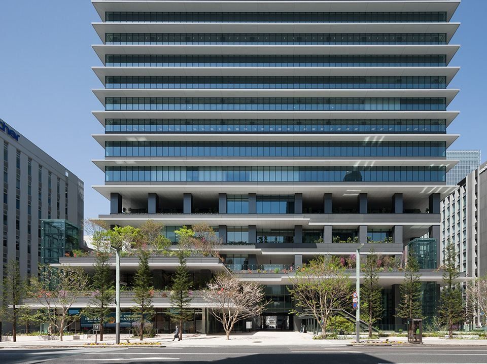 A tall glass office tower with horizontal bands rising above terraced, tree-planted lower floors