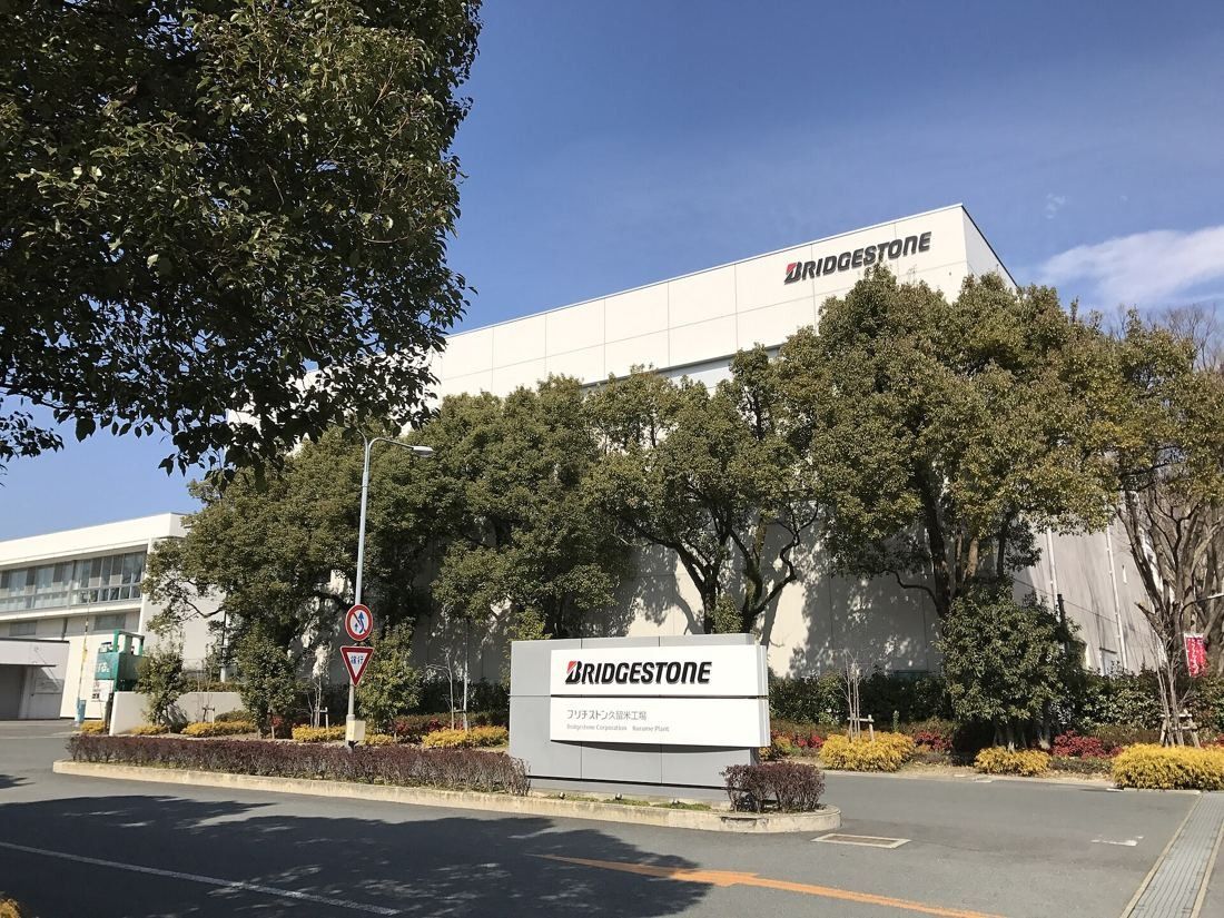A white factory building behind trees with a company sign at the gate