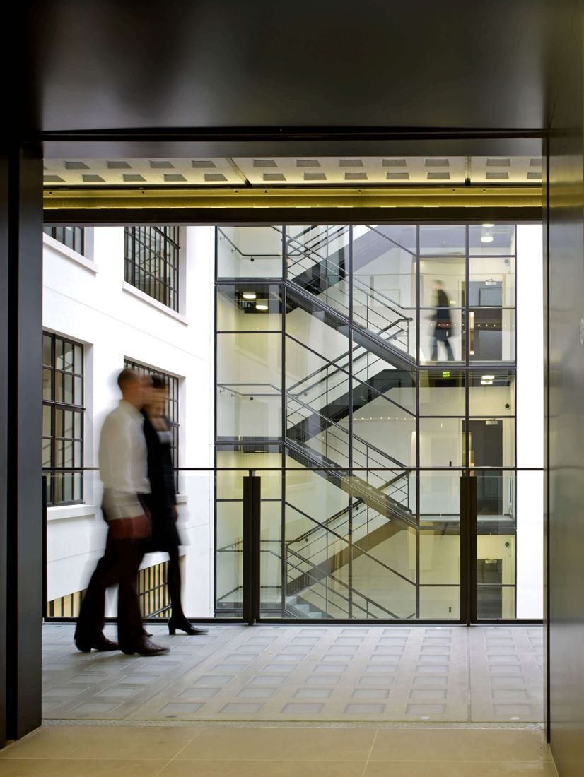 Burberry: Blurred figures walking along a bridge past a glass-enclosed staircase in a white atrium