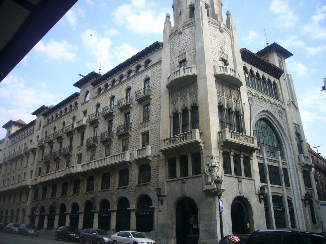 Stone building with a tall pointed tower and a large arched window on a city street