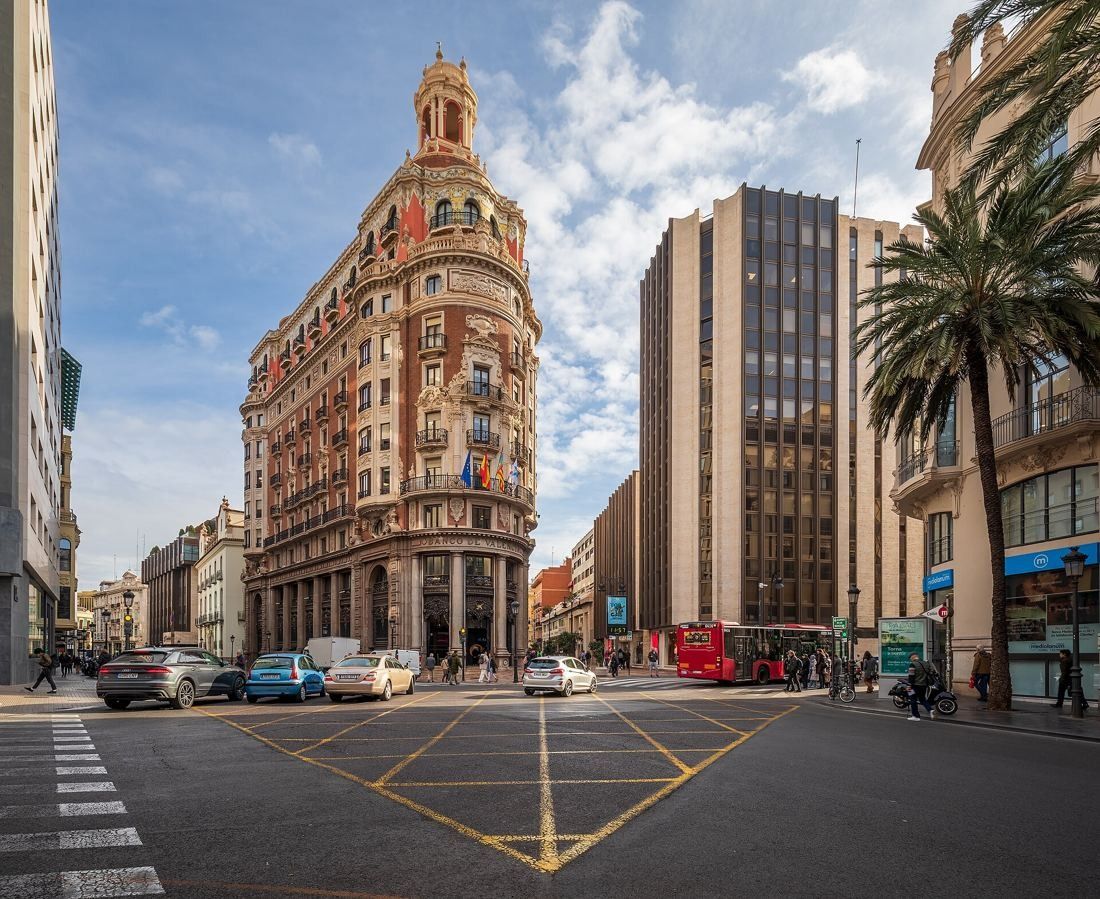 Wide view of a crossroads with an ornate brick-and-stone corner building topped by a domed lantern, a dark glass office block beside it