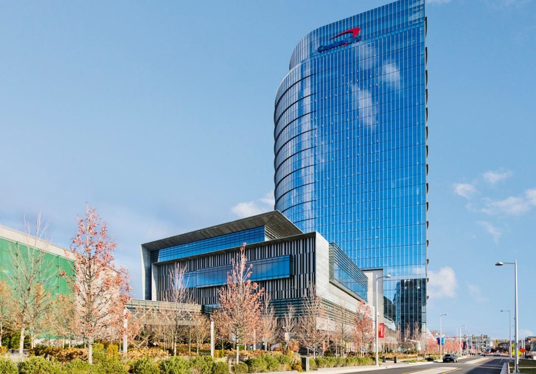Tall curved blue glass tower rising behind a lower glass building and young trees