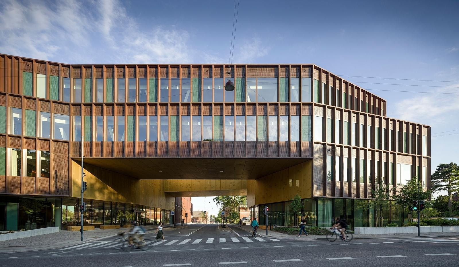 Copper-coloured building with vertical slats raised over a glazed, open ground floor facing a street