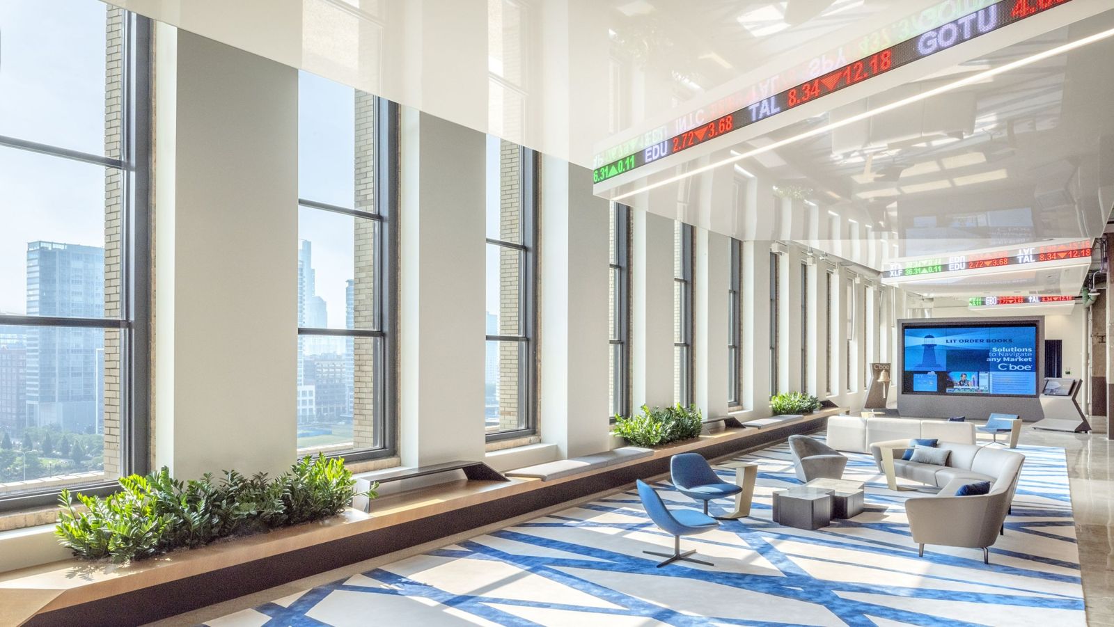 A long lounge under a white ceiling with scrolling stock tickers and tall windows over Chicago