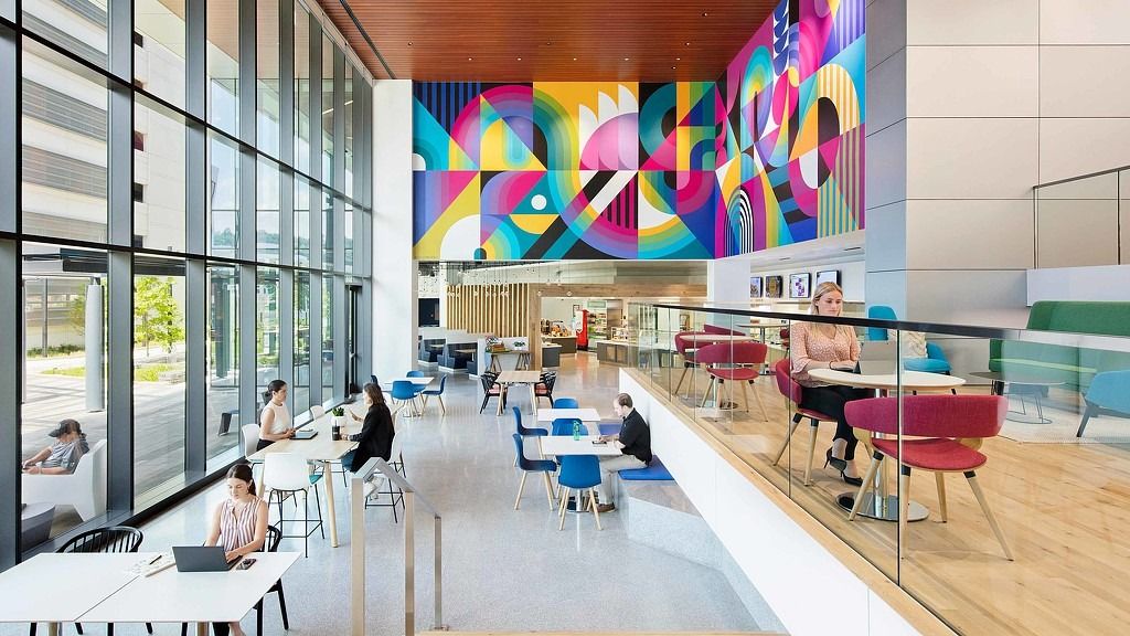A bright café floor with colourful murals, a long counter and windows onto a plaza