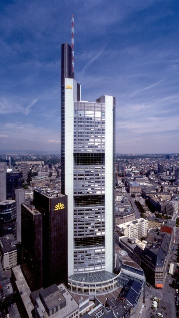 Commerzbank: Aerial view of a white tower with a red and white antenna mast above a dark neighbouring high-rise and the city