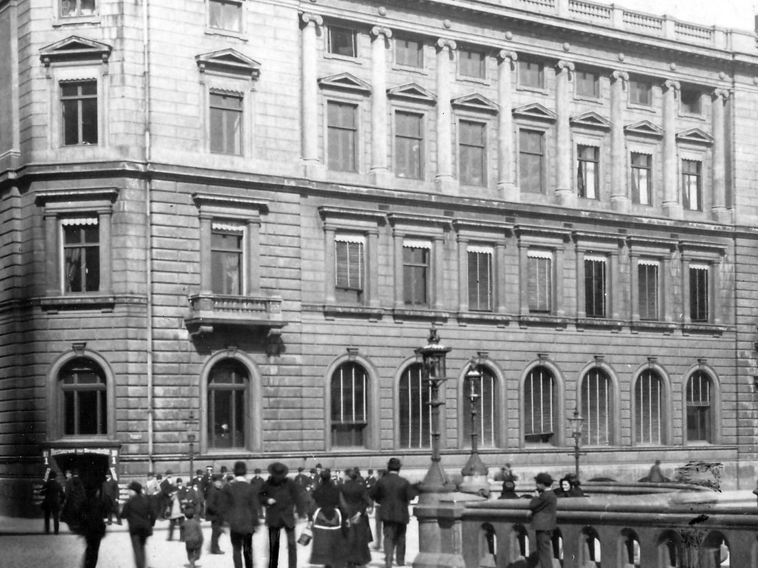 Black and white photo of a large stone bank building beside a statue on a bridge, with people on the pavement
