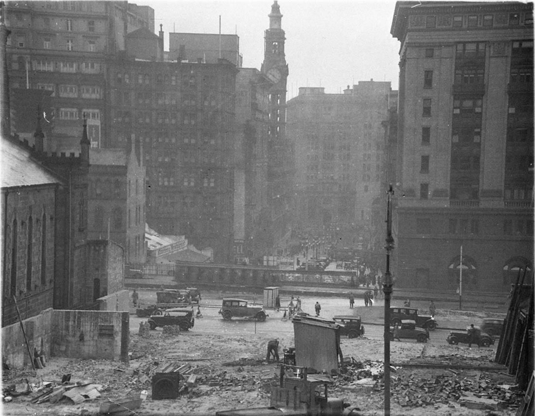 Black-and-white view along a wide street under construction with a tall bank building on the right