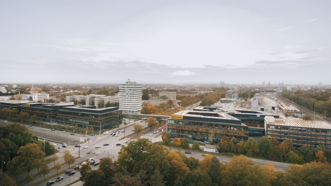 Continental: Aerial view of low glass office buildings on both sides of a wide road, linked by a bridge, with autumn trees and the city behind