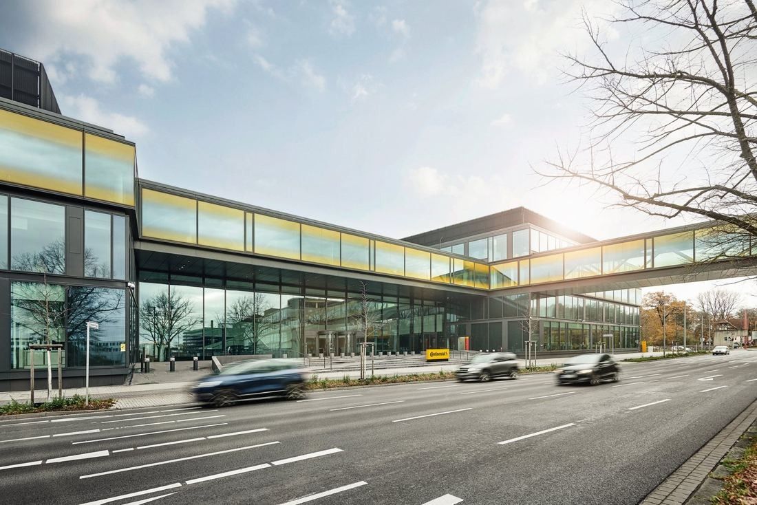 Glass bridge with a yellow band spanning a multi-lane road between two glass office buildings, cars passing below