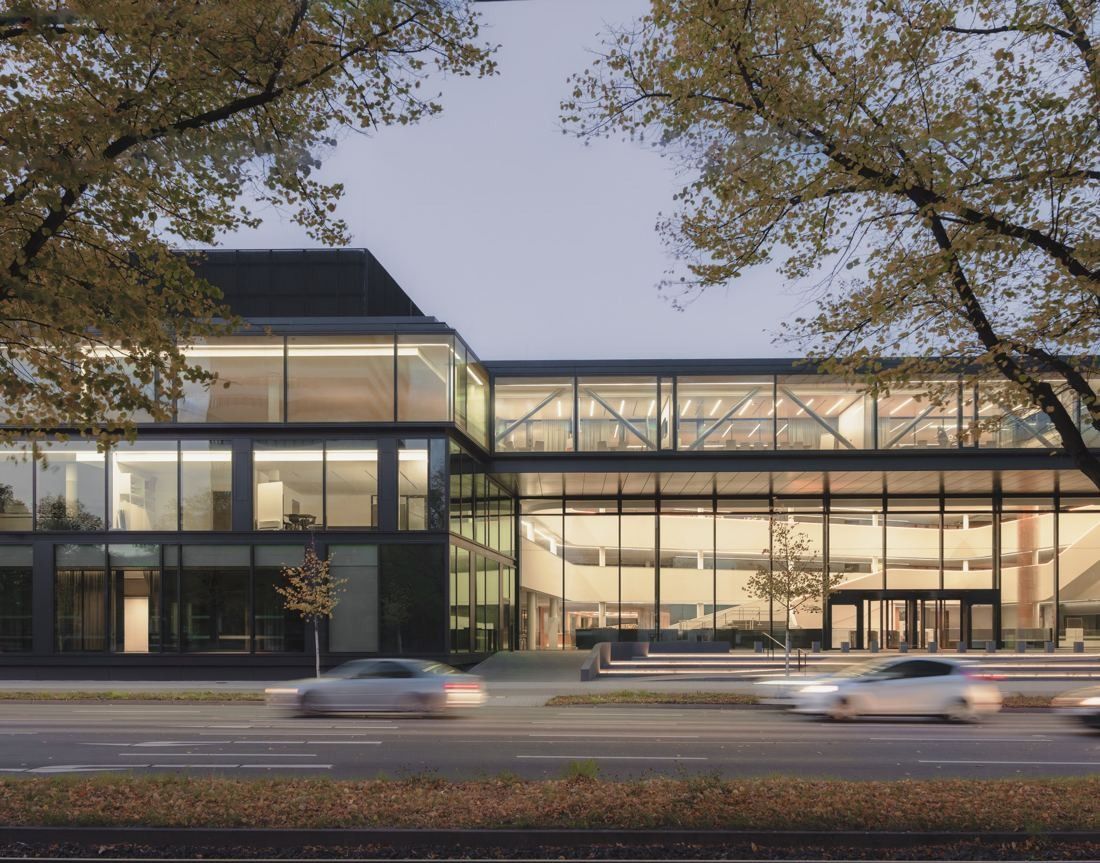 Continental: Glass office building at dusk with lit floors and a glazed walkway, traffic blurred in front