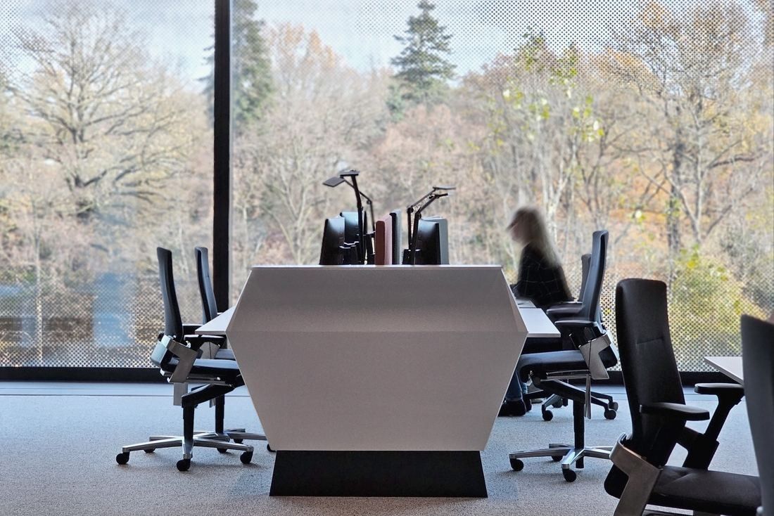 Continental: White angular desk with monitors and black office chairs in front of a perforated window with trees outside