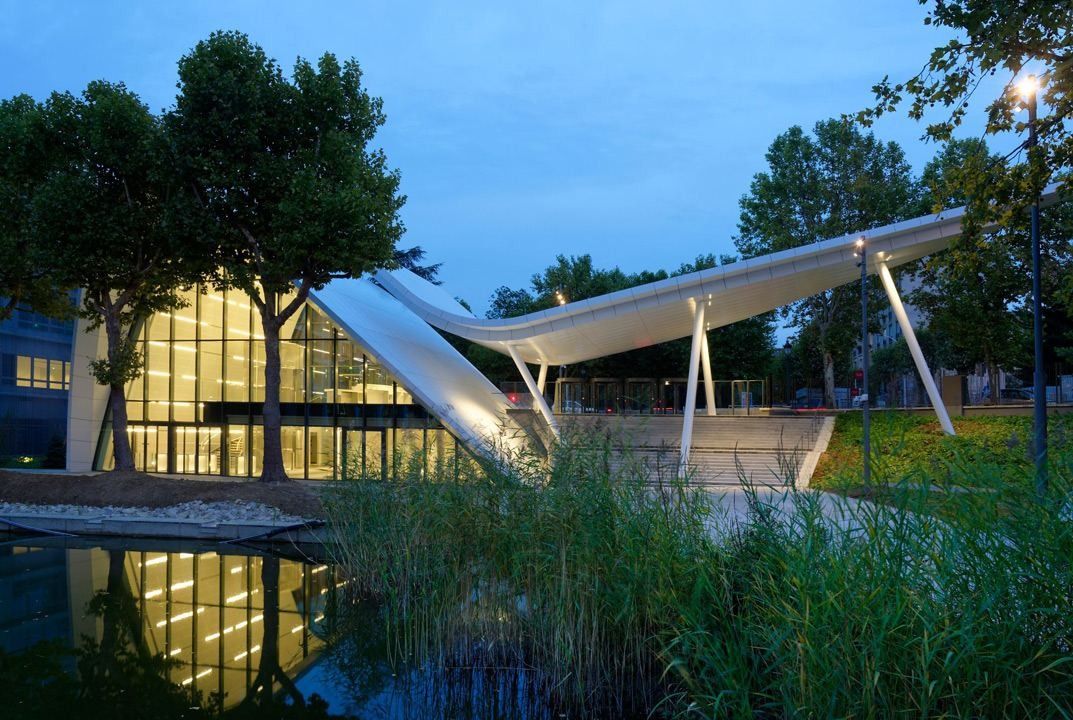 A white curved canopy roof over a glazed pavilion, reflected in a planted pond at dusk
