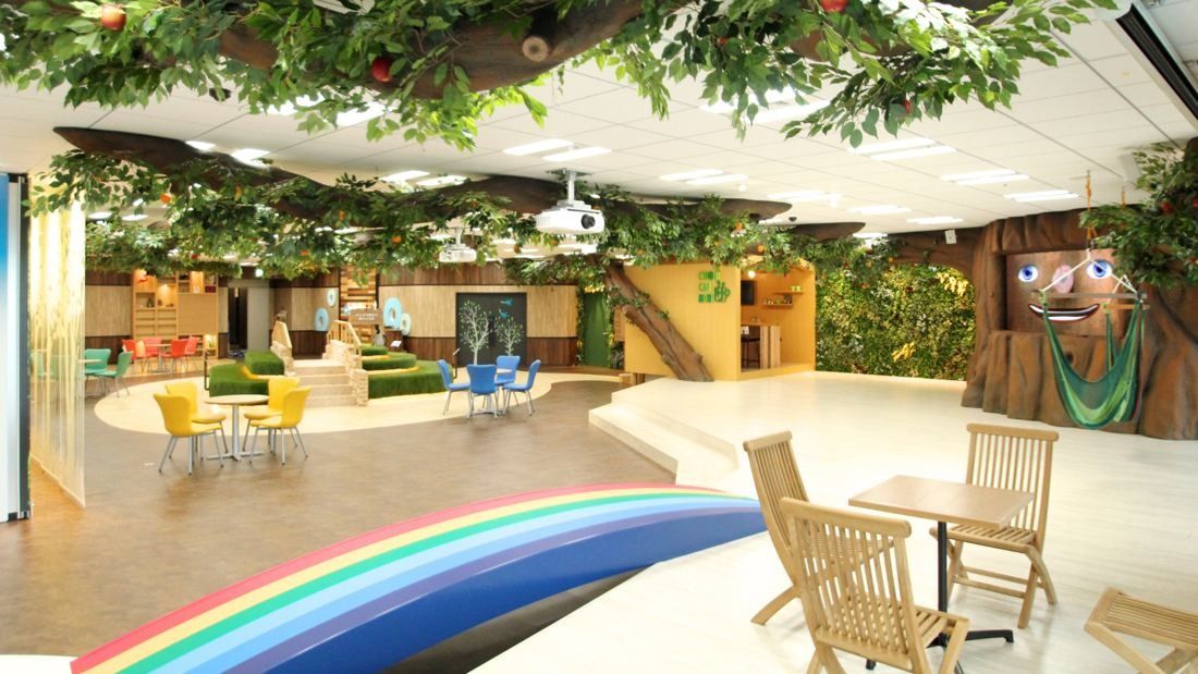 Cybozu: Entrance area under a canopy of artificial trees, with a rainbow bridge, wooden chairs and a tree-shaped figure