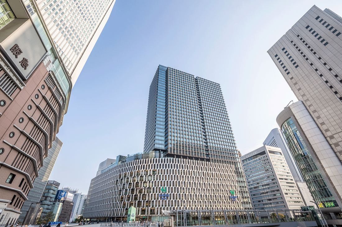 A tall glass tower rising above a curved, patterned lower block among city high-rises, seen from below on a clear day