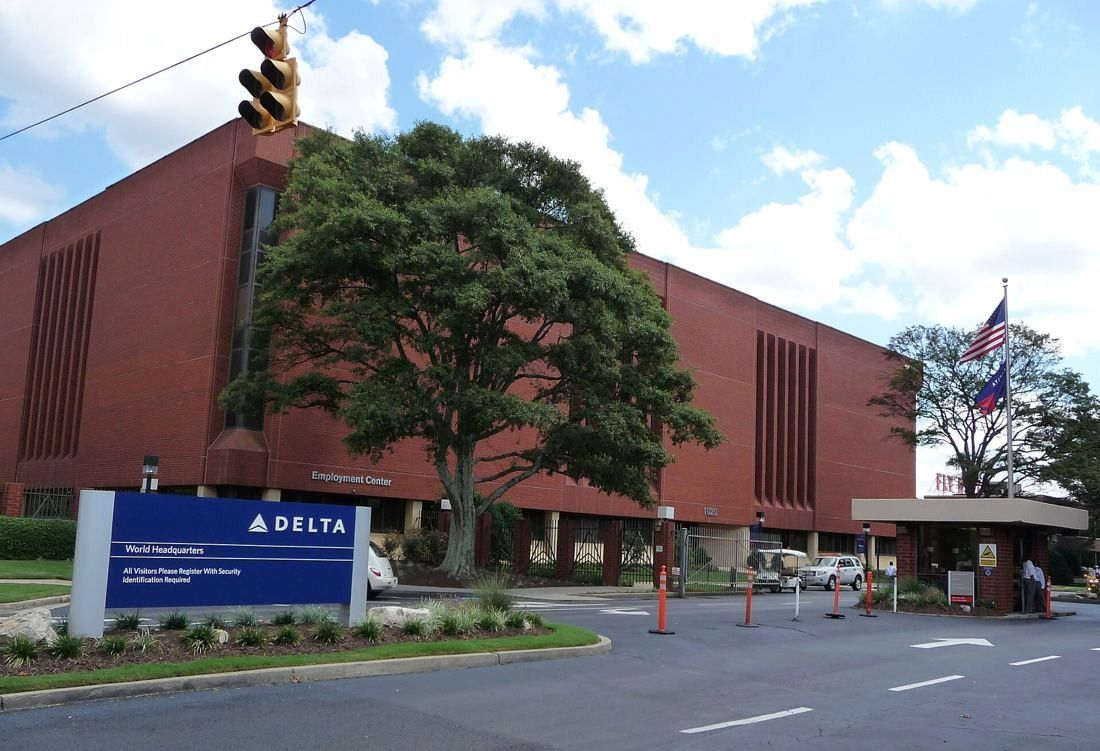Long red-brick office building with vertical window bands behind a large tree, a blue Delta sign and a guard booth at the entrance drive