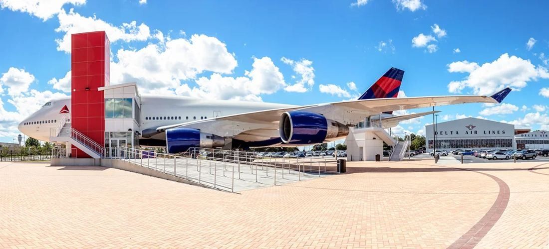 Delta Air Lines: Panorama of a Boeing 747 in Delta livery on display outdoors beside a red stair tower, with hangar buildings in the distance