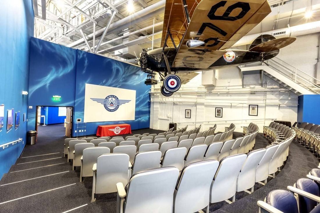 Delta Air Lines: Theater with rows of white seats inside a hangar, blue walls, a screen and a vintage plane hanging overhead