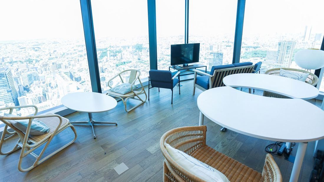 DeNA: Round white tables and wooden chairs beside tall windows over the city