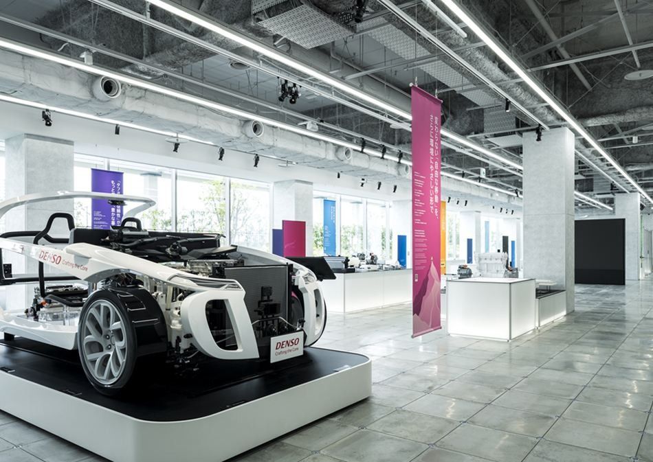 Denso: Bright hall with exposed ceiling ducts, a white car chassis on a platform and display cases beyond