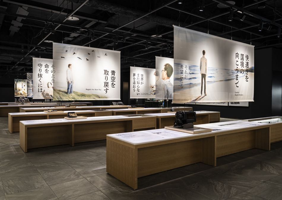 Denso: Dim gallery with rows of wooden display tables beneath large hanging printed banners
