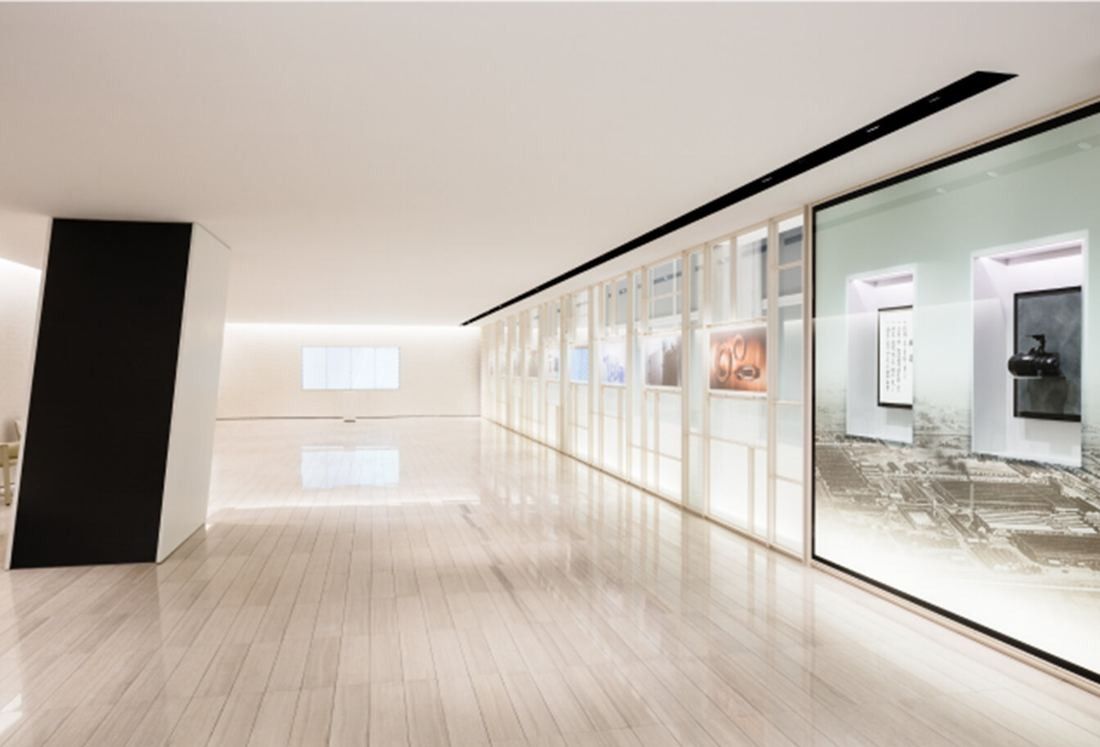 Denso: Long pale corridor with a black slanted column and a glass wall of framed exhibits