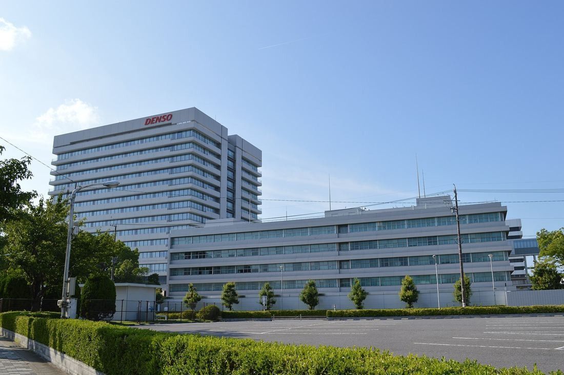 Denso: Low white office building in front of a taller white tower, with a parking lot and young trees