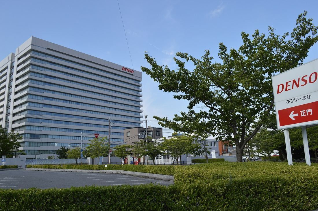 Denso: Tall white office tower behind trees, with a red and white company sign pointing to the main gate