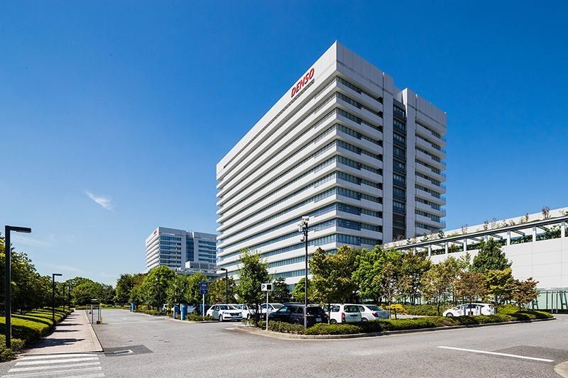 Denso: White high-rise office tower beside a tree-lined road and parking lot under a clear blue sky