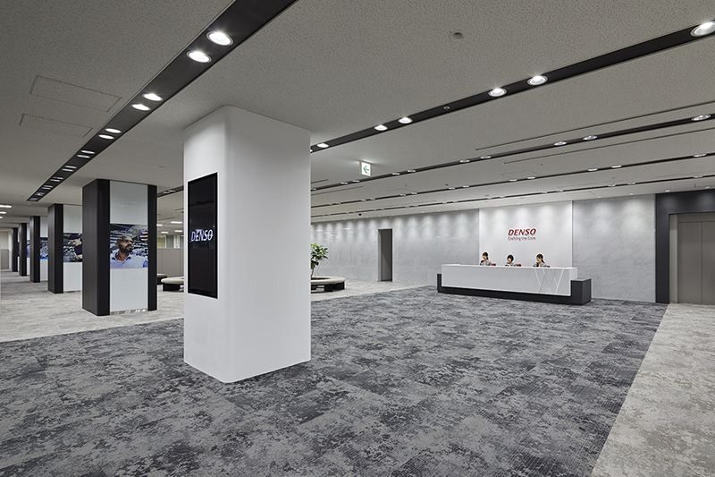 Denso: Wide reception lobby with grey patterned carpet, white columns with screens and a reception desk in the distance