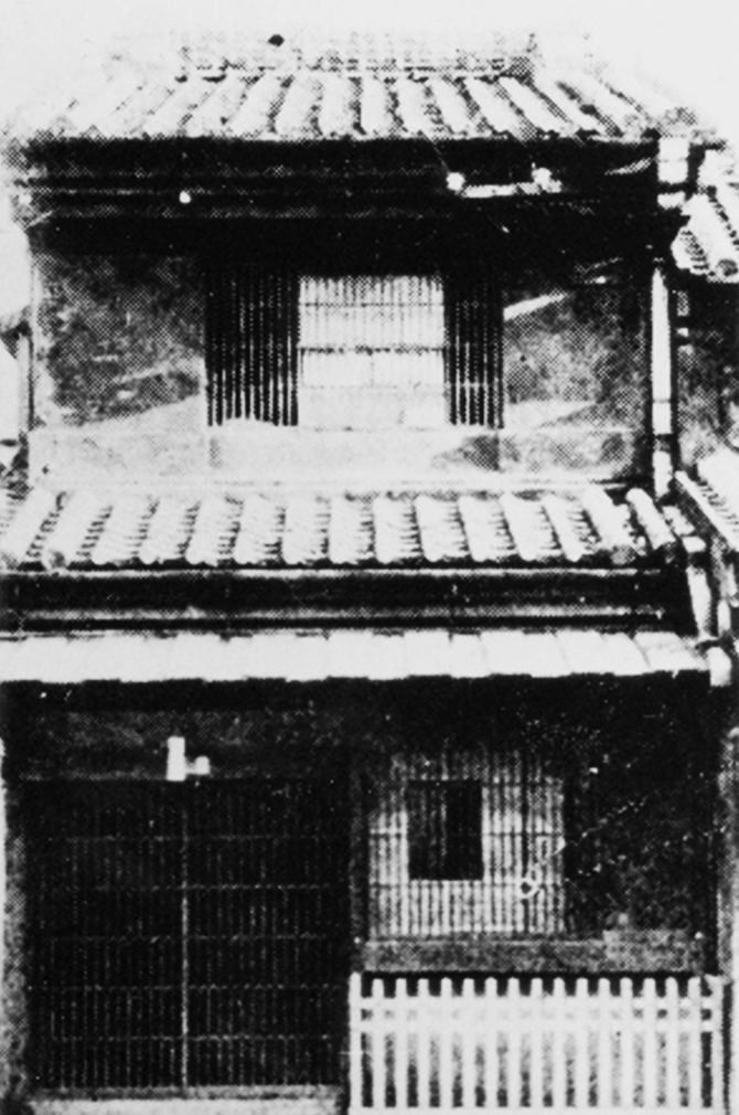 Old black-and-white photo of a narrow two-storey wooden house with a tiled roof and lattice front