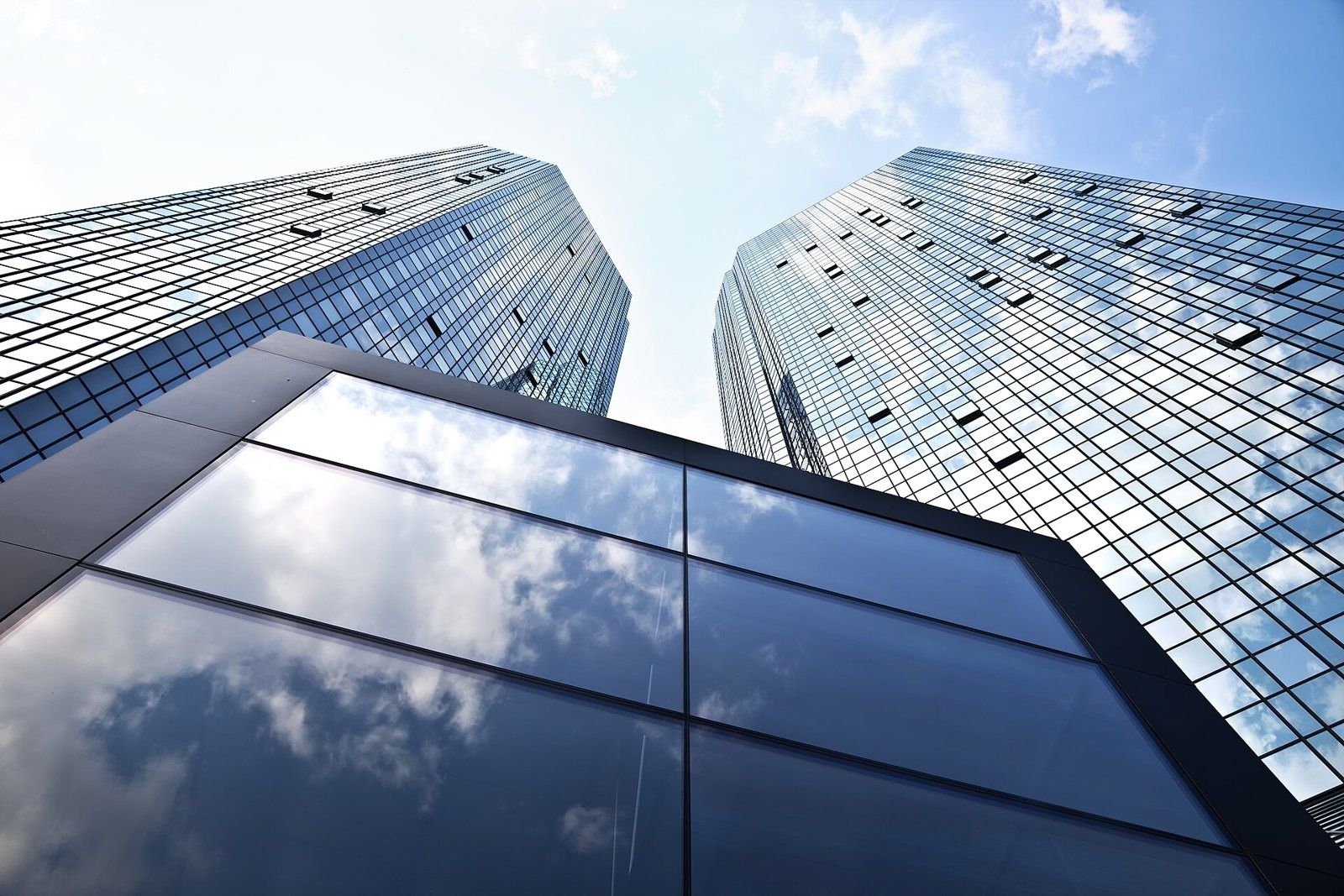 Looking up at two glass towers rising above a glass entrance canopy reflecting clouds