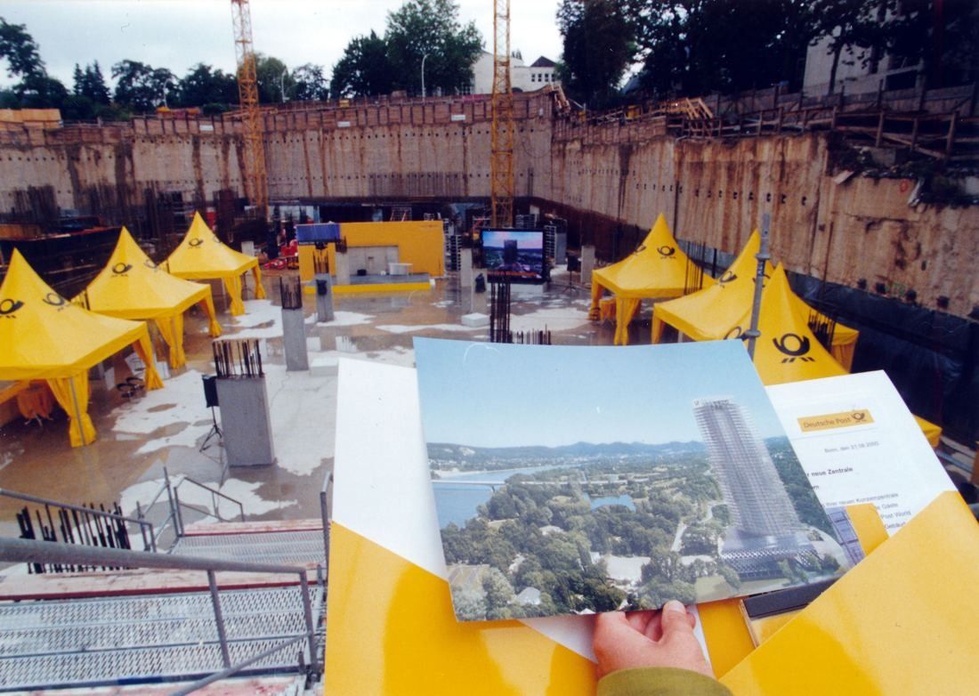 Excavated construction pit with yellow tents, and a hand holding a brochure showing the planned tower
