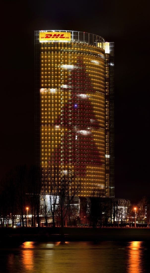 DHL Group: Tower at night with yellow and red lights forming a shape on the façade, above a river