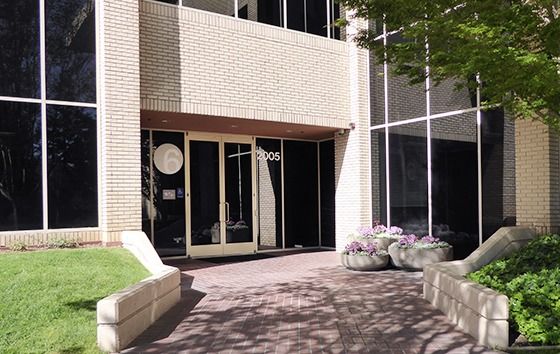 Entrance of a low beige office building with dark windows, a paved path and shrubs