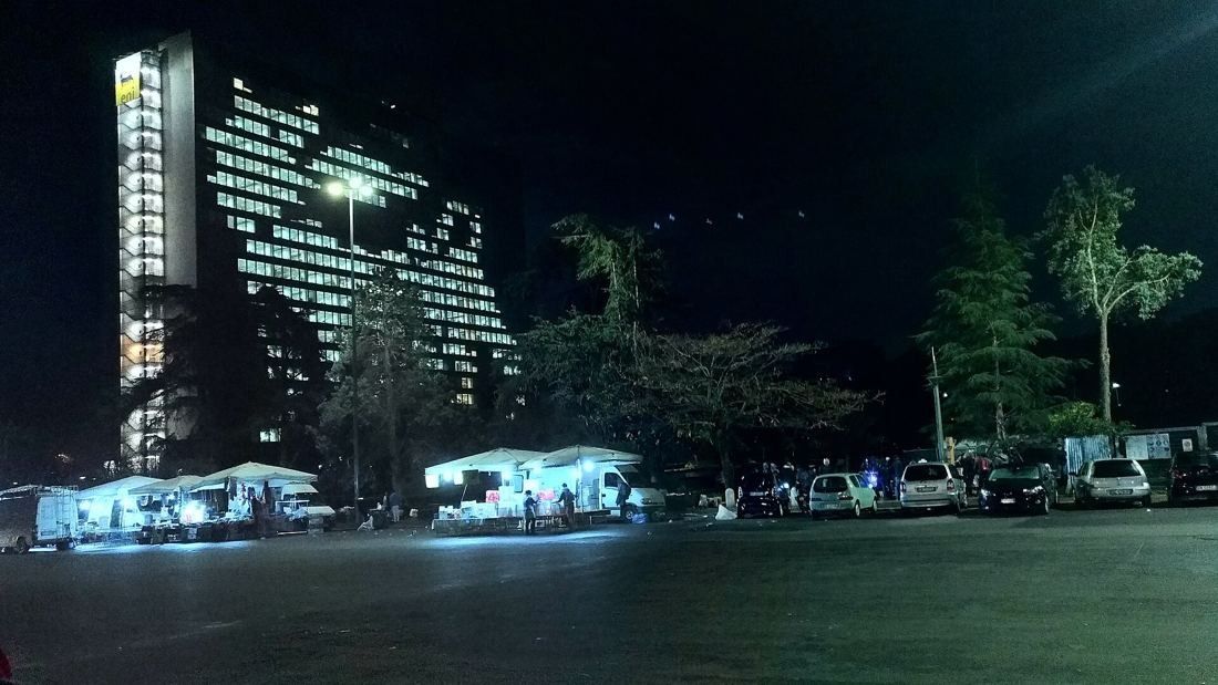 Eni: A glass tower with lit windows at night above trees and a parking area
