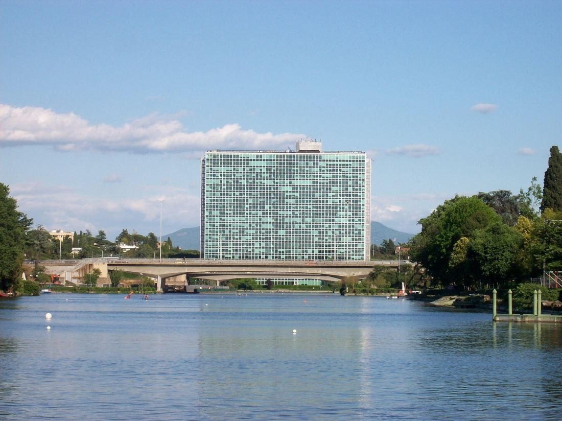 A tall glass office slab rising beyond a calm lake, with a low bridge and trees in front