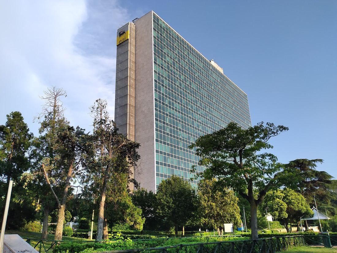 Eni: A tall glass tower with a stone end wall seen above trees in a park