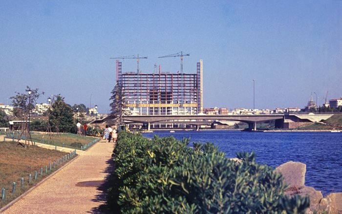 A tall office building under construction with cranes, seen across a lake in Rome