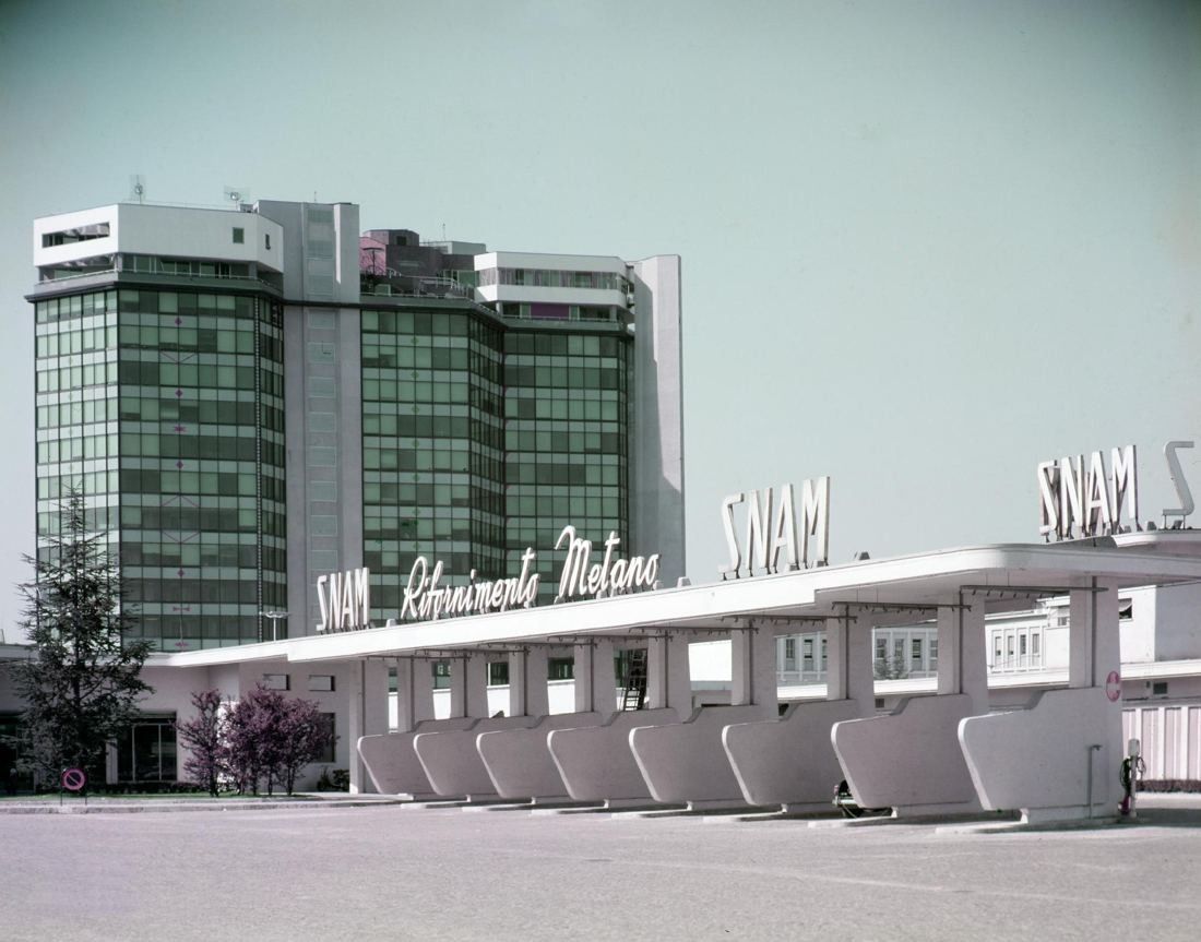Black-and-white style photo of a glass office tower behind a long canopy with SNAM lettering