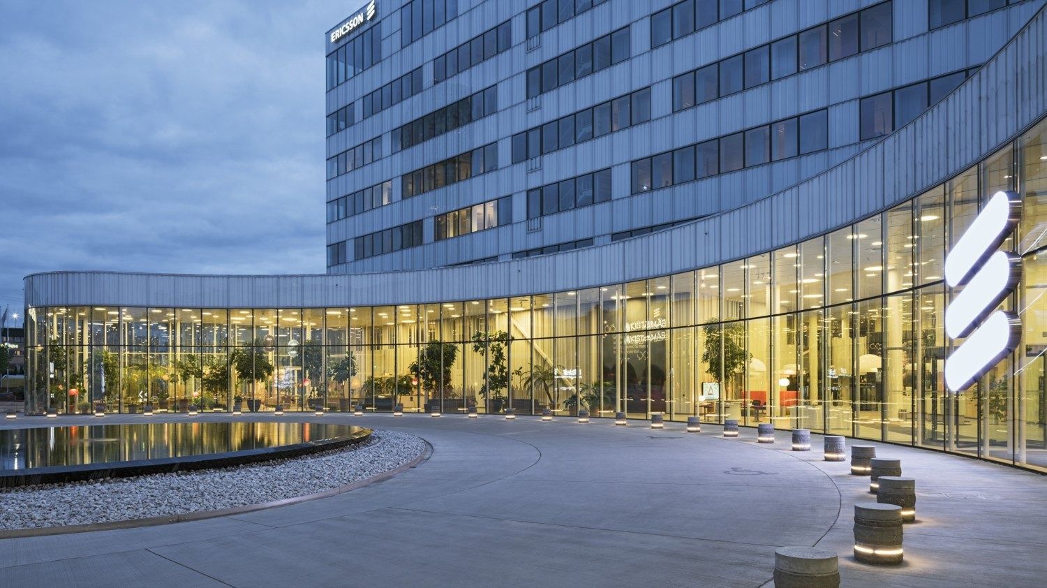 Ericsson: Curved glass ground floor with a lit logo beside a reflecting pond at dusk