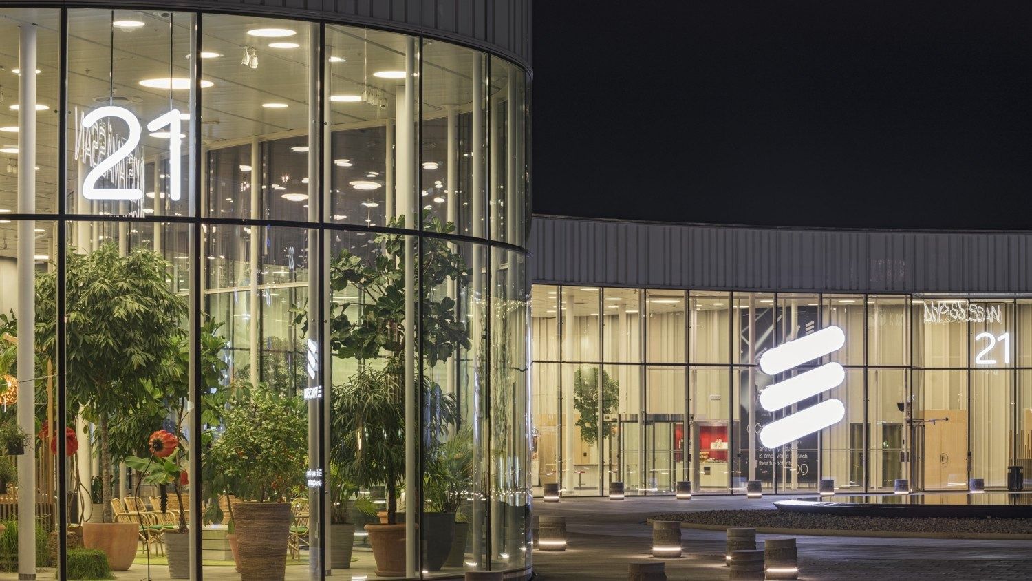 Ericsson: Glass entrance with indoor trees and a lit logo, seen at night