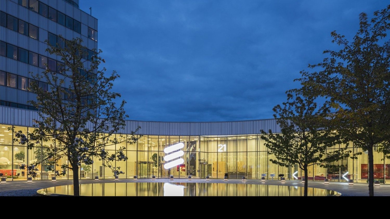 Ericsson: Lit glass pavilion behind a pond, framed by two trees at dusk