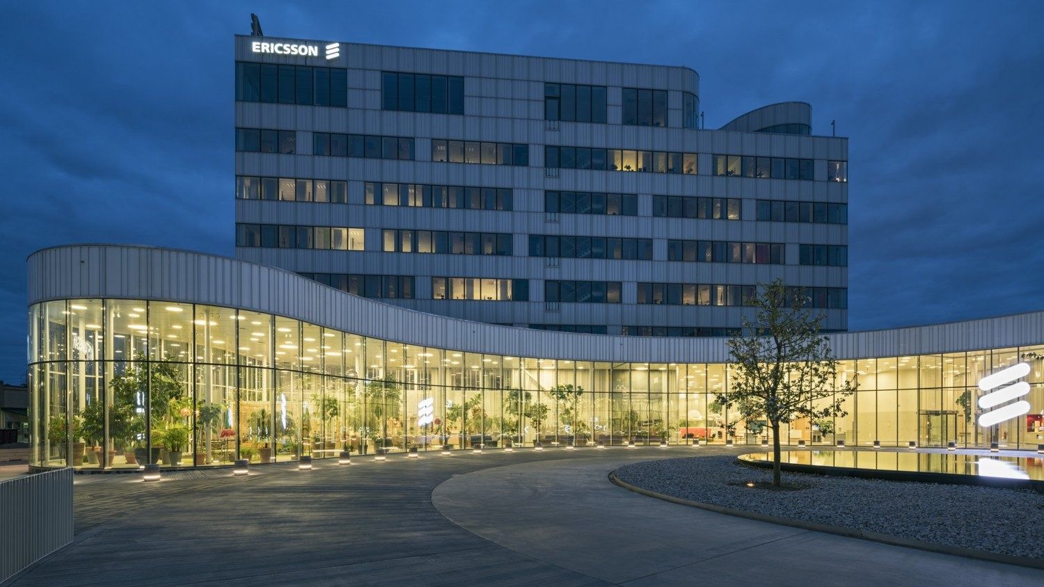 Office building at dusk with a lit, curved glass ground-floor pavilion and forecourt