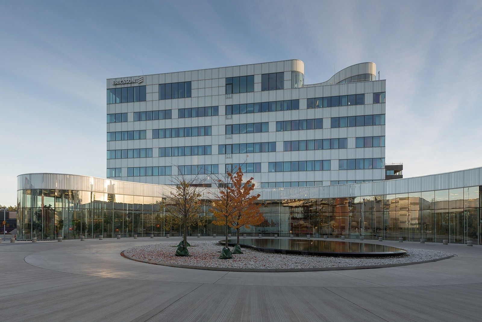Ericsson: White office building behind a curved glass base and round pond, autumn trees