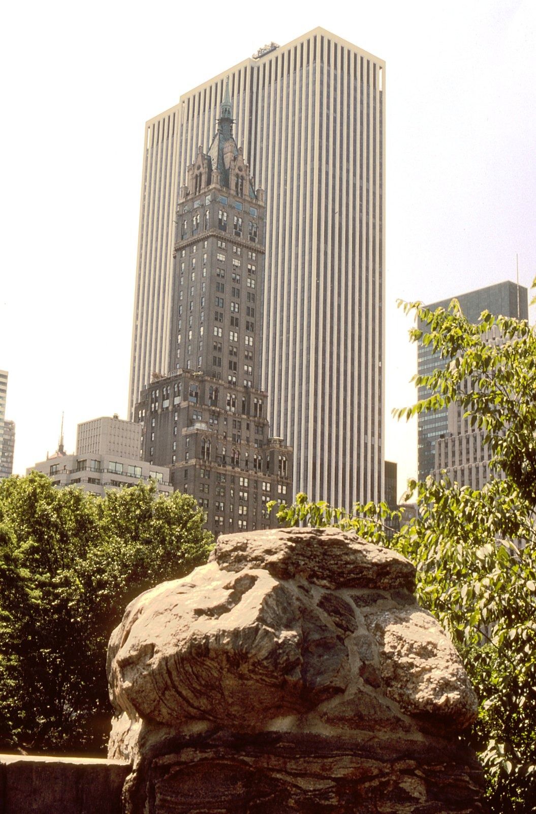 The Estée Lauder Companies: Ornate brown tower in front of a taller white-striped skyscraper, rock and trees in the foreground